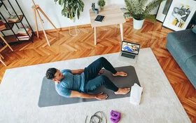 Das Bild zeigt einen jungen Mann im Wohnzimmer auf einer Yogamatte. Neben ihm steht ein Laptop, auf dem ein Workout-Video eingeschaltet ist.