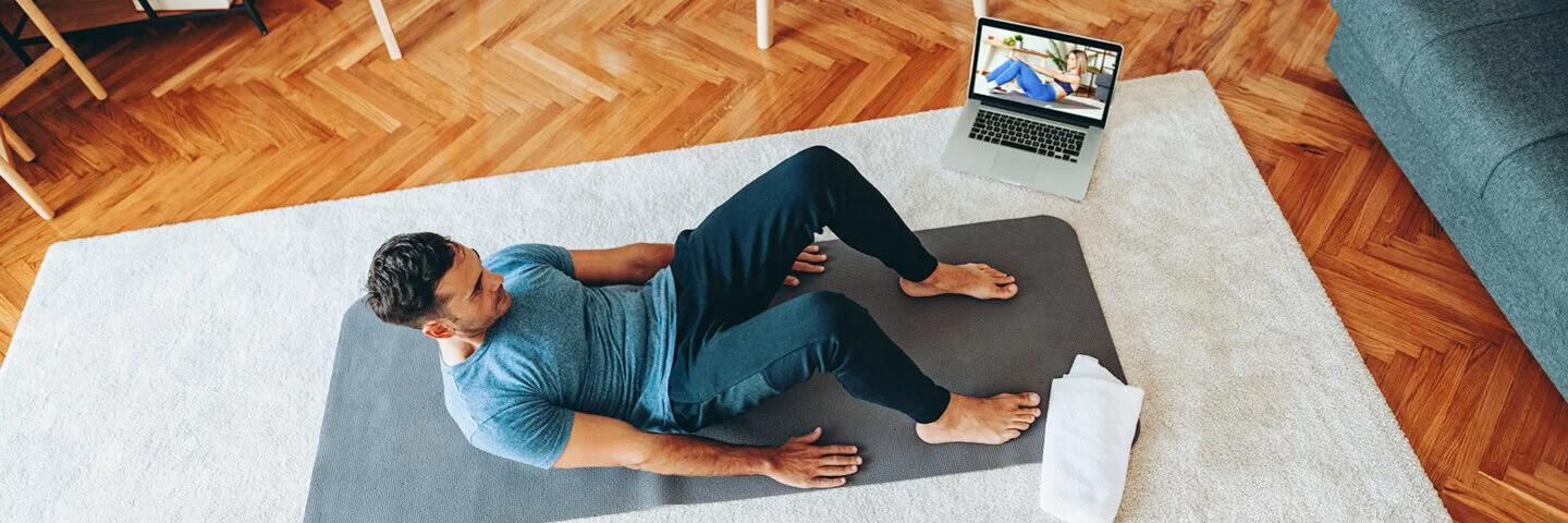 Das Bild zeigt einen jungen Mann im Wohnzimmer auf einer Yogamatte. Neben ihm steht ein Laptop, auf dem ein Workout-Video eingeschaltet ist.