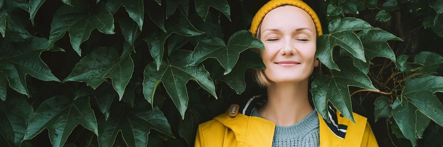 Ein Frau steht mit geschlossenen Augen in einem gelben Regenmantel vor einen Blätterwand.