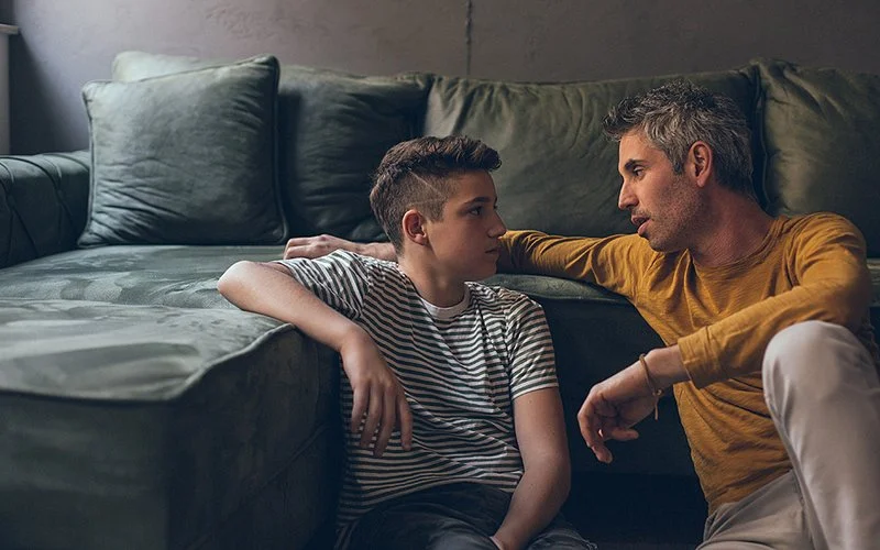 Ein Vater und sein Sohn sitzen gemeinsam auf dem Fußboden im Wohnzimmer und unterhalten sich.