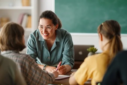 Eine junge Lehrerin mit Brille spricht mit einer Gruppe von Schülern im Klassenzimmer. Sie lächelt freundlich und lehnt sich über einen Tisch, auf dem Hefte und Stifte liegen. Im Hintergrund ist eine grüne Tafel und ein Regal mit Büchern zu sehen.