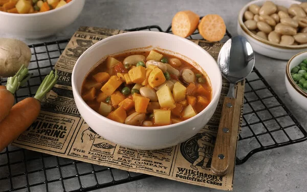 Weiße Schale mit veganem Gemüsetopf mit Süßkartoffeln. Die Schale steht auf einem silbernen Drahtgitter auf grauem Untergrund. Rund herum sind Kartoffeln, Möhren, Erbsen, dicke Bohnen und Süßkartoffeln drapiert.