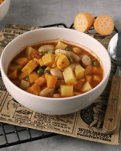 Weiße Schale mit veganem Gemüsetopf mit Süßkartoffeln. Die Schale steht auf einem silbernen Drahtgitter auf grauem Untergrund. Rund herum sind Kartoffeln, Möhren, Erbsen, dicke Bohnen und Süßkartoffeln drapiert.