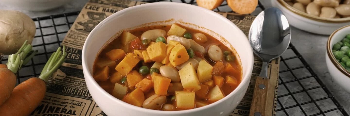 Weiße Schale mit veganem Gemüsetopf mit Süßkartoffeln. Die Schale steht auf einem silbernen Drahtgitter auf grauem Untergrund. Rund herum sind Kartoffeln, Möhren, Erbsen, dicke Bohnen und Süßkartoffeln drapiert.