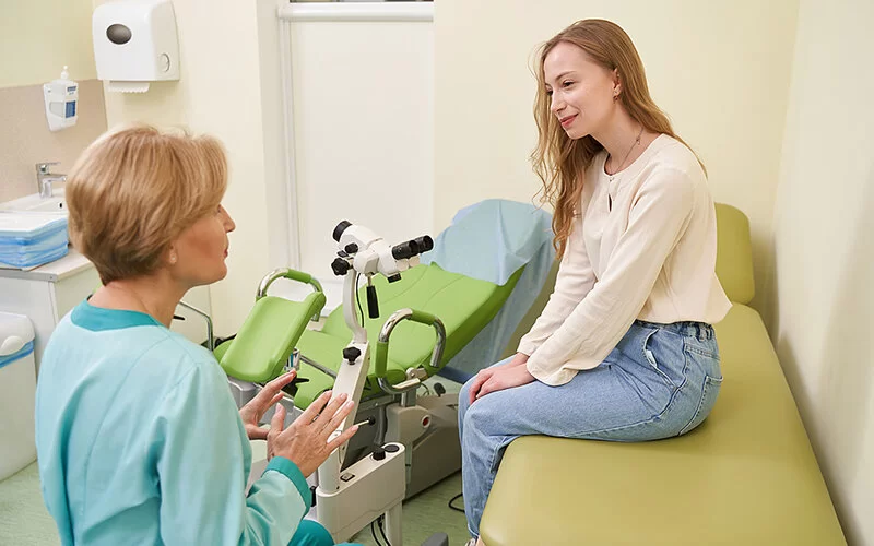 Eine Patientin sitzt angezogen auf einer Behandlungsliege. Auf einem Hocker vor ihr sitzt die Frauenärztin und erklärt der Patientin gendersensible Sprache – zum Beispiel, dass Vulvalippen ein passenderes Wort für Schamlippen ist.