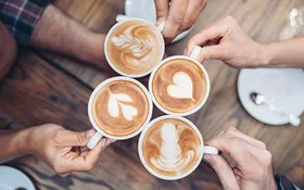 Ansicht von vier Händen, die jeweils eine mit Kaffee gefüllte Tasse halten.
