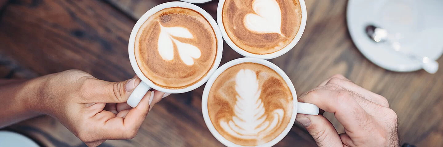 Ansicht von vier Händen, die jeweils eine mit Kaffee gefüllte Tasse halten.