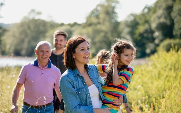 Bei sonnigem Wetter geht eine fröhlich lächelnde Familie durch eine natürliche, grüne Umgebung. Eine Frau hält ein junges Mädchen auf dem Arm. Im Hintergrund sind etwas verschwommen ein älterer Mann, ein mittelalter Mann und eine weitere Person zu sehen.
