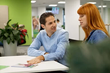 Ein Mann sitzt an einem Schreibtisch und sieht eine Frau freundlich an, die links neben ihm sitzt.