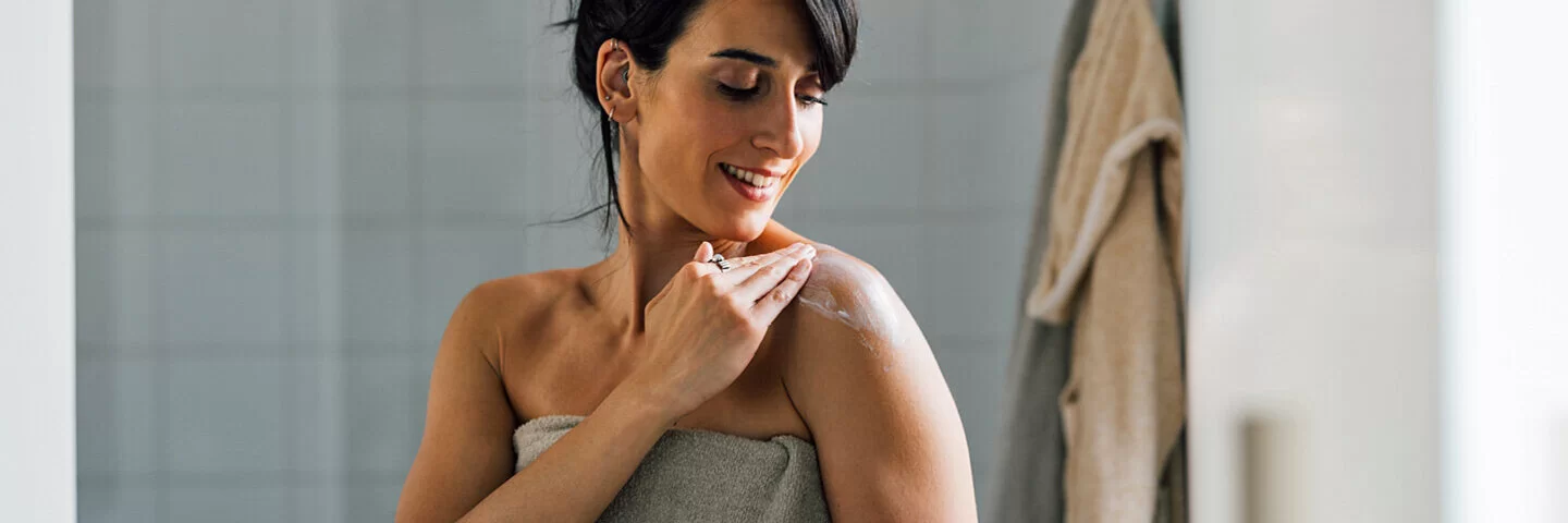 Eine Frau mit Handtuch um den Körper cremt sich nach der Dusche ein.