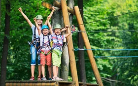 Drei fröhliche Kinder mit Helmen und Kletterharnischen sind in einem Hochseilgarten zu sehen.