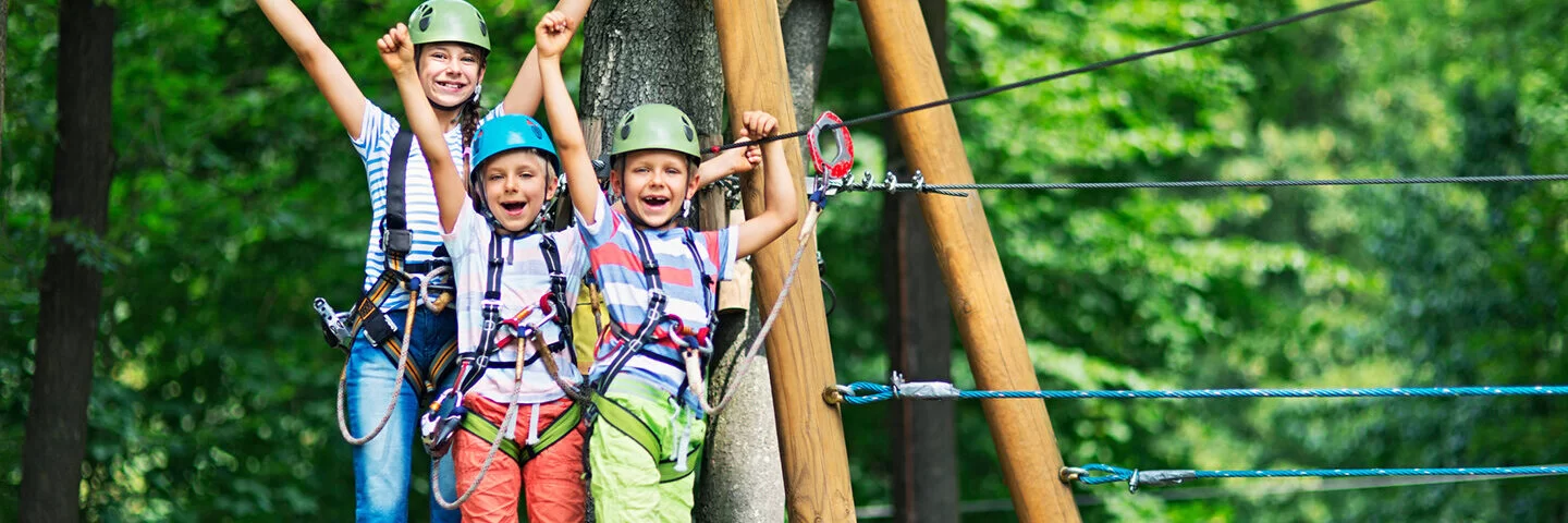 Drei fröhliche Kinder mit Helmen und Kletterharnischen sind in einem Hochseilgarten zu sehen.