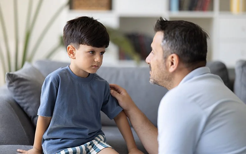 Ein Junge sitzt auf dem Sofa, ein Mann kniet vor ihm und legt dem Jungen eine Hand auf die Schulter.