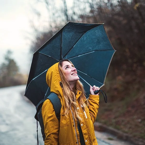 Eine junge Frau steht im Regen, über sich hat sie einen Regenschirm gespannt. Sie schaut zuversichtlich in den Himmel.