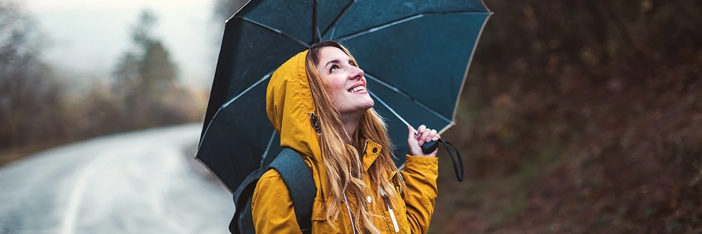 Eine junge Frau steht im Regen, über sich hat sie einen Regenschirm gespannt. Sie schaut zuversichtlich in den Himmel.