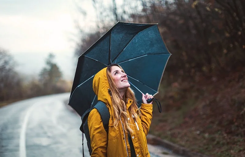 Eine junge Frau steht im Regen, über sich hat sie einen Regenschirm gespannt. Sie schaut zuversichtlich in den Himmel.