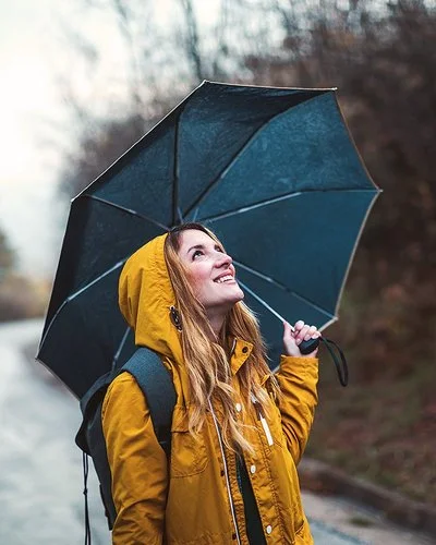 Eine junge Frau steht im Regen, über sich hat sie einen Regenschirm gespannt. Sie schaut zuversichtlich in den Himmel.