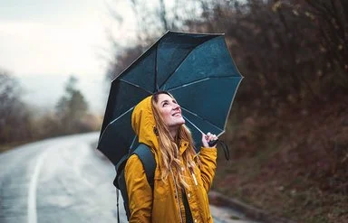 Eine junge Frau steht im Regen, über sich hat sie einen Regenschirm gespannt. Sie schaut zuversichtlich in den Himmel.