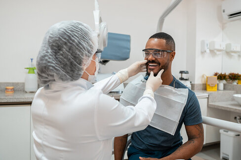 Ein Zahnarzt tastet den Kiefer eines Patienten ab. Beide tragen Schutzkleidung, um eine Infektion mit dem Coronavirus in der AOK-Zahnklinik zu verhindern.