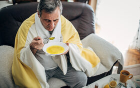 Ein Mann sitzt auf dem Sofa und löffelt eine warme Suppe.