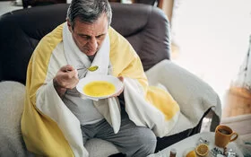 Ein Mann sitzt auf dem Sofa und löffelt eine warme Suppe.