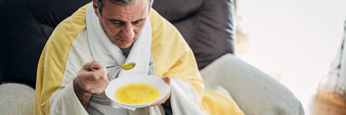 Ein Mann sitzt auf dem Sofa und löffelt eine warme Suppe.