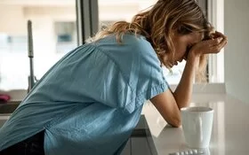 Eine Frau hat starke Kopfschmerzen und fasst sich an den Kopf, neben ihr auf dem Tisch liegen Kopfschmerztabletten.