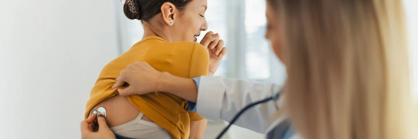 Eine Ärztin hört bei einer Patientin mit einem Stethoskop die Atmung ab. Mit der rechten Hand schiebt sie das Shirt der Patientin nach oben, während sie mit der linken Hand den Kopf des Stethoskops im oberen Rückenbereich hält.