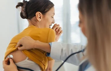 Eine Ärztin hört bei einer Patientin mit einem Stethoskop die Atmung ab. Mit der rechten Hand schiebt sie das Shirt der Patientin nach oben, w ährend sie mit der linken Hand den Kopf des Stethoskops im oberen Rückenbereich hält.