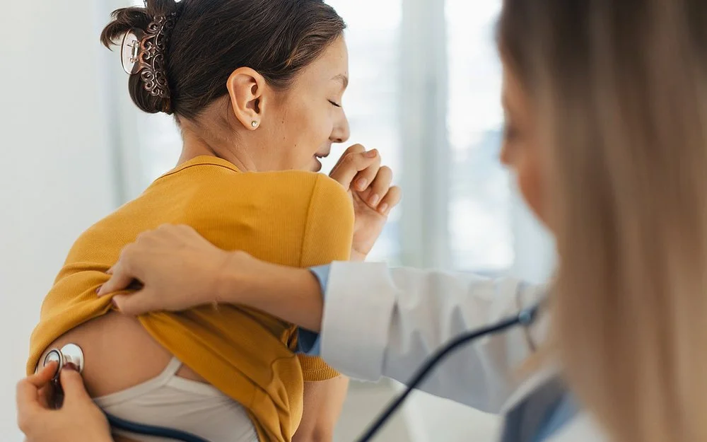 Eine Ärztin hört bei einer Patientin mit einem Stethoskop die Atmung ab. Mit der rechten Hand schiebt sie das Shirt der Patientin nach oben, während sie mit der linken Hand den Kopf des Stethoskops im oberen Rückenbereich hält.