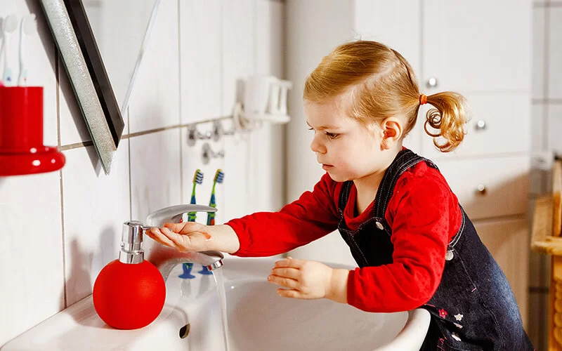 Ein ungefähr fünfjähriges Mädchen steht auf einem Hocker am Waschbecken in einem Badezimmer. Der Wasserhahn läuft und sie nimmt Seife aus einem Spender, um sich die Hände zu waschen.