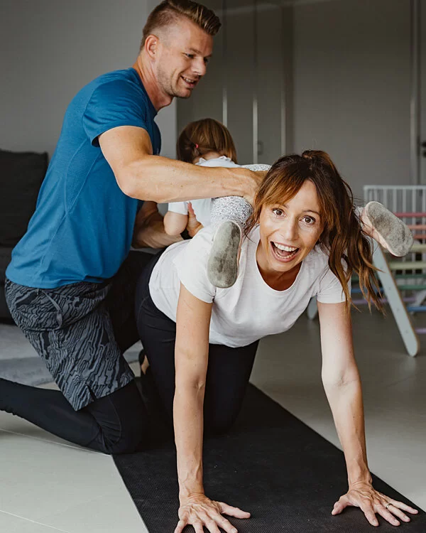 Daniela macht Yoga, während ihre Tochter auf ihrem Rücken klettert und Mann Steffen sie hält.