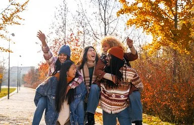 Fünf junge Erwachsenen laufen durch einen herbstlichen, sonnigen Park. Zwei Personen tragen jeweils eine andere auf dem Rücken, während alle lachend und ausgelassen wirken. Bunte Herbstbäume sind im Hintergrund zu sehen.
