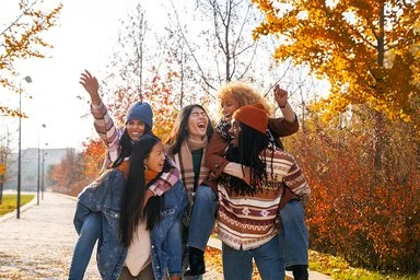 Fünf junge Erwachsenen laufen durch einen herbstlichen, sonnigen Park. Zwei Personen tragen jeweils eine andere auf dem Rücken, während alle lachend und ausgelassen wirken. Bunte Herbstbäume sind im Hintergrund zu sehen.