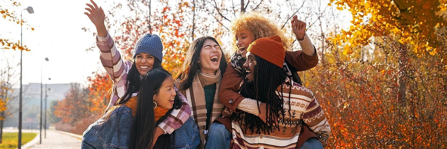 Fünf junge Erwachsenen laufen durch einen herbstlichen, sonnigen Park. Zwei Personen tragen jeweils eine andere auf dem Rücken, während alle lachend und ausgelassen wirken. Bunte Herbstbäume sind im Hintergrund zu sehen.