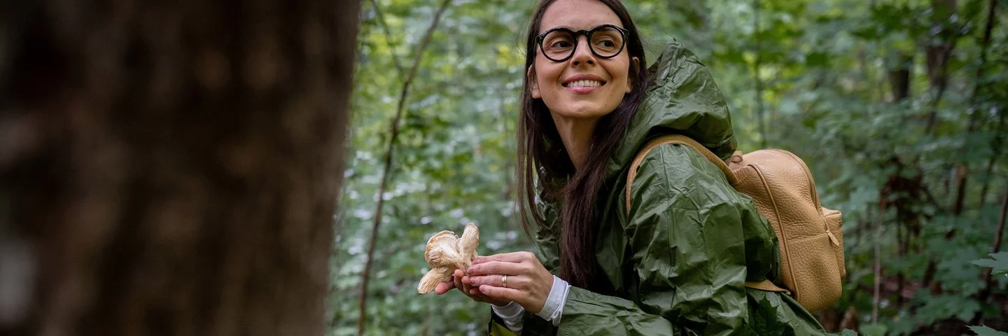 Eine junge lachende Frau steht im Wald und hält einen großen Pilz in einer Hand.