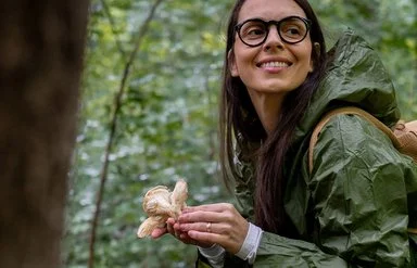 Eine junge lachende Frau steht im Wald und hält einen großen Pilz in einer Hand.