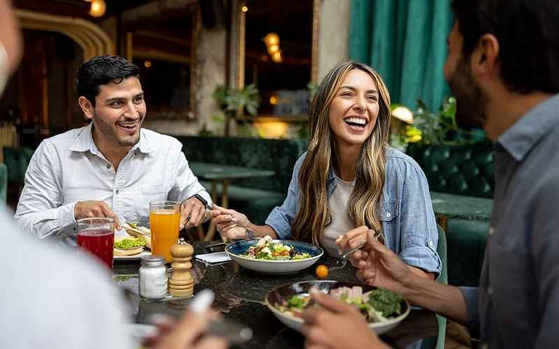 Vier Menschen sitzen gemeinsam in einem Restaurant und essen Salate. Sie lachen und unterhalten sich während der Mahlzeit.