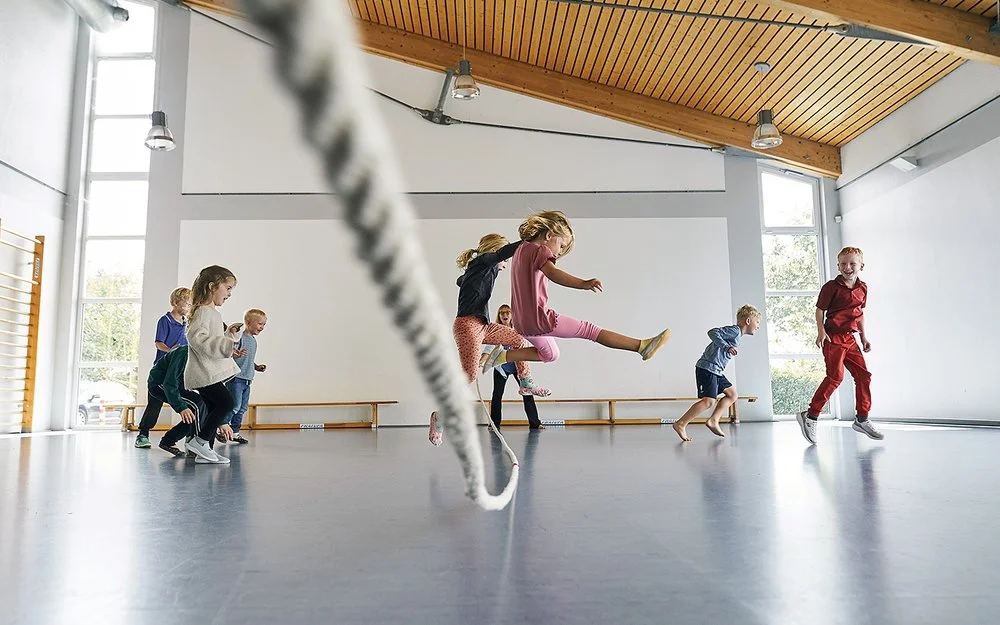 In einer durch Tageslicht erhellten Turnhalle springen mehrere Kinder in Sportkleidung über ein langes Tau. Ein Mädchen in einem rosa T-Shirt springt zentral in die Luft. Andere Kinder rennen oder springen freudig links und rechts daneben.