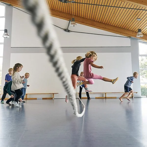 In einer durch Tageslicht erhellten Turnhalle springen mehrere Kinder in Sportkleidung über ein langes Tau. Ein Mädchen in einem rosa T-Shirt springt zentral in die Luft. Andere Kinder rennen oder springen freudig links und rechts daneben.