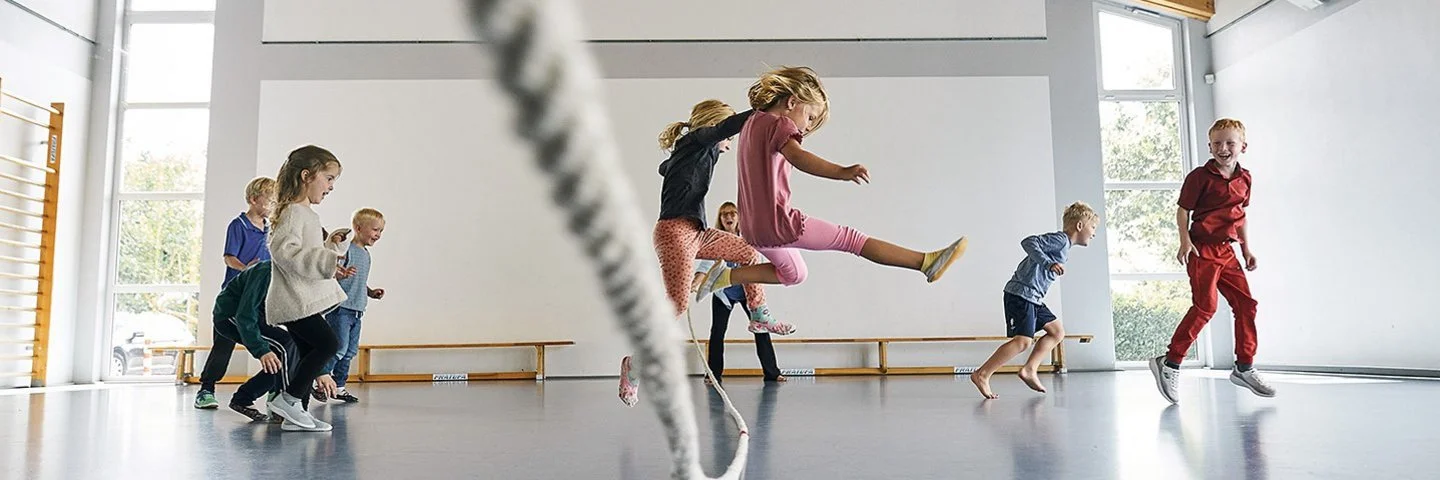 In einer durch Tageslicht erhellten Turnhalle springen mehrere Kinder in Sportkleidung über ein langes Tau. Ein Mädchen in einem rosa T-Shirt springt zentral in die Luft. Andere Kinder rennen oder springen freudig links und rechts daneben.