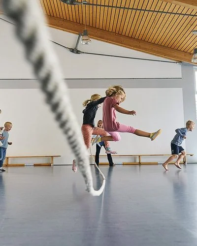 In einer durch Tageslicht erhellten Turnhalle springen mehrere Kinder in Sportkleidung über ein langes Tau. Ein Mädchen in einem rosa T-Shirt springt zentral in die Luft. Andere Kinder rennen oder springen freudig links und rechts daneben.