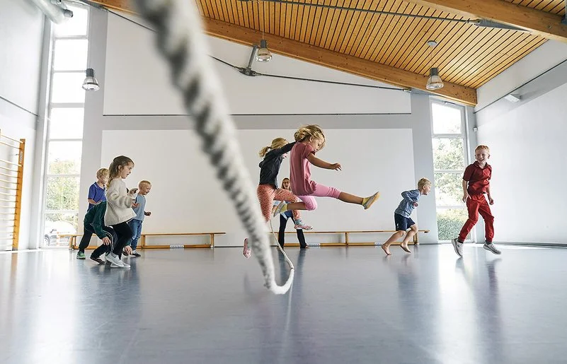 In einer durch Tageslicht erhellten Turnhalle springen mehrere Kinder in Sportkleidung über ein langes Tau. Ein Mädchen in einem rosa T-Shirt springt zentral in die Luft. Andere Kinder rennen oder springen freudig links und rechts daneben.