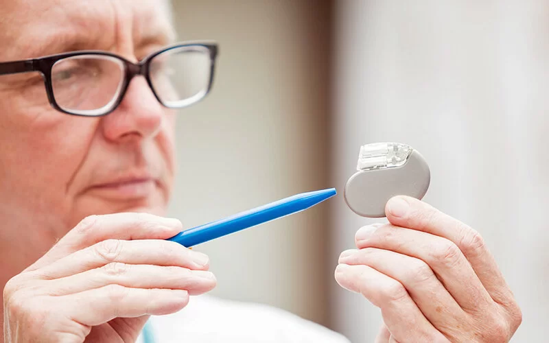 Ein älterer Arzt hält einen Herzschrittmacher in der Hand und erklärt mit einem Stift das Gerät.