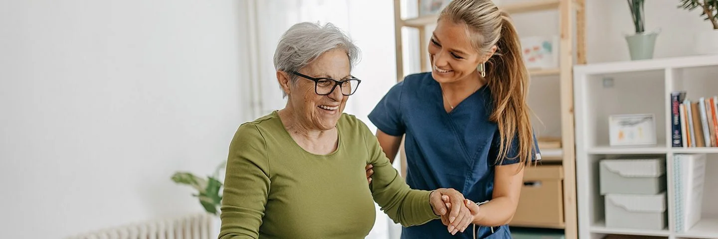 Eine Pflegerin hilft einer alten Frau mit Gehstock beim Laufen. Die Pflegerin stützt sie unter dem Arm. Beide befinden sich in einem hellen Wohnzimmer.