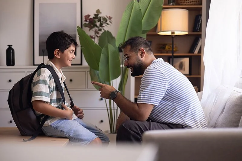 Ein Mann mit Brille und gestreiftem T-Shirt sitzt auf einem Sofa und spricht lächelnd mit einem Jungen mit Rucksack, der auf einem Stuhl sitzt