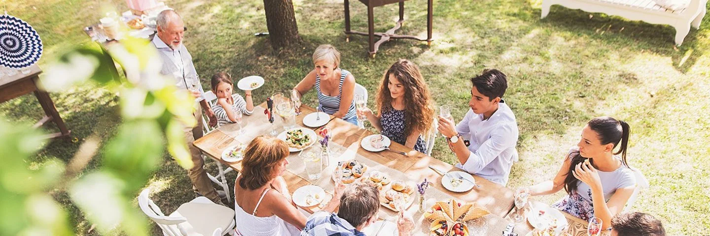 Eine große, mehrgenerationelle Familie sitzt an einem langen Tisch im dekorierten Garten und unterhält sich während des Essens.