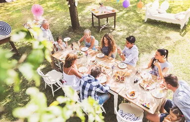 Eine große, mehrgenerationelle Familie sitzt an einem langen Tisch im dekorierten Garten und unterhält sich während des Essens.