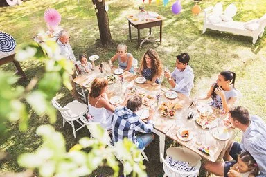 Eine große, mehrgenerationelle Familie sitzt an einem langen Tisch im dekorierten Garten und unterhält sich während des Essens.