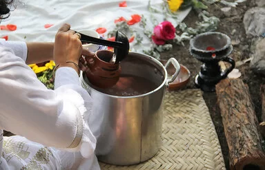 Eine Frau schöpft mit einer Kelle eine Flüssigkeit aus einem Topf in eine Tasse. Im Hintergrund sieht man Blumen.